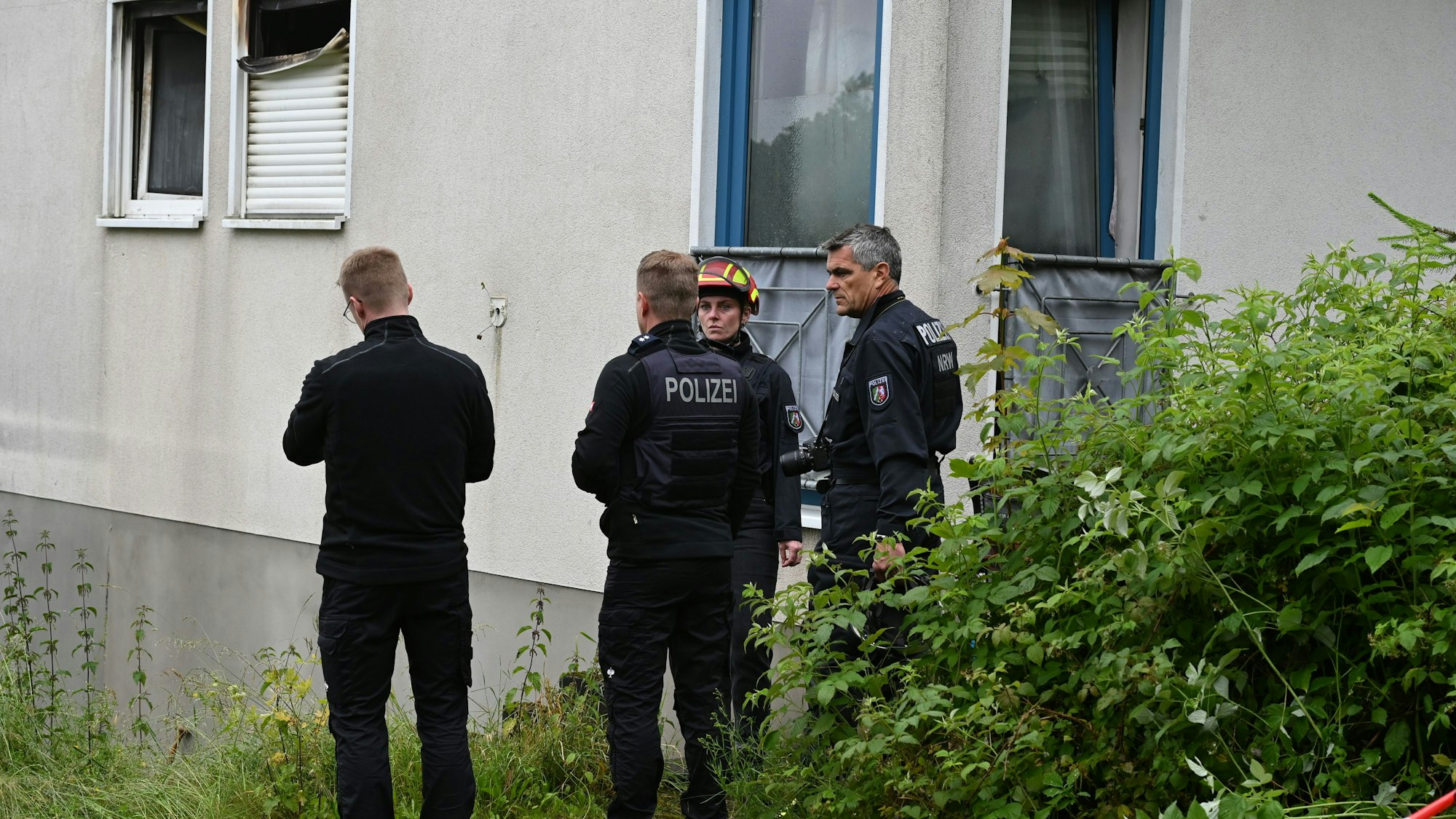 Drei Männer und eine Frau stehen vor einer Hauswand. Ein Rollladen ist durch Flammen zerstört worden.