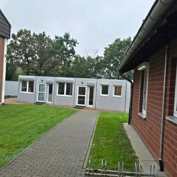 Zwischen zwei Gebäuden der Kita-Houverath wurde ein Container platziert. Ein gepflasterter Weg führt dorthin, flankiert von Rasenflächen.