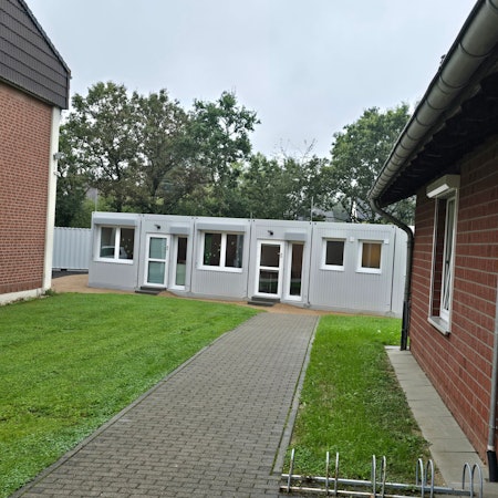 Zwischen zwei Gebäuden der Kita-Houverath wurde ein Container platziert. Ein gepflasterter Weg führt dorthin, flankiert von Rasenflächen.