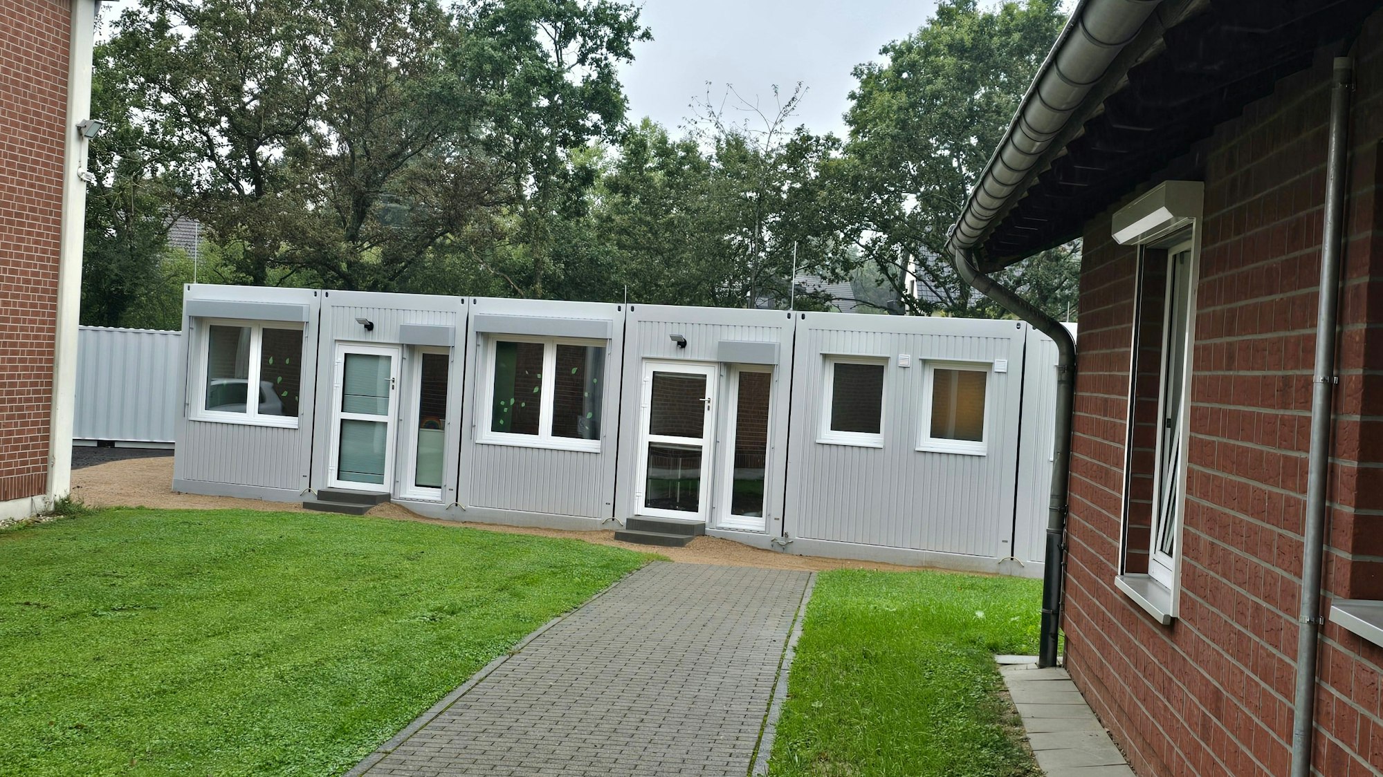 Zwischen zwei Gebäuden der Kita-Houverath wurde ein Container platziert. Ein gepflasterter Weg führt dorthin, flankiert von Rasenflächen.