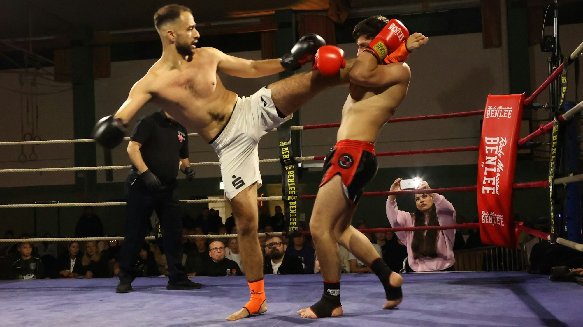 Ein Kickboxer hat den Fuß des Gegners auf der Schulter liegen und greift mit dem Arm danach. Im Hintergrund fotografiert eine Frau mit dem Handy.