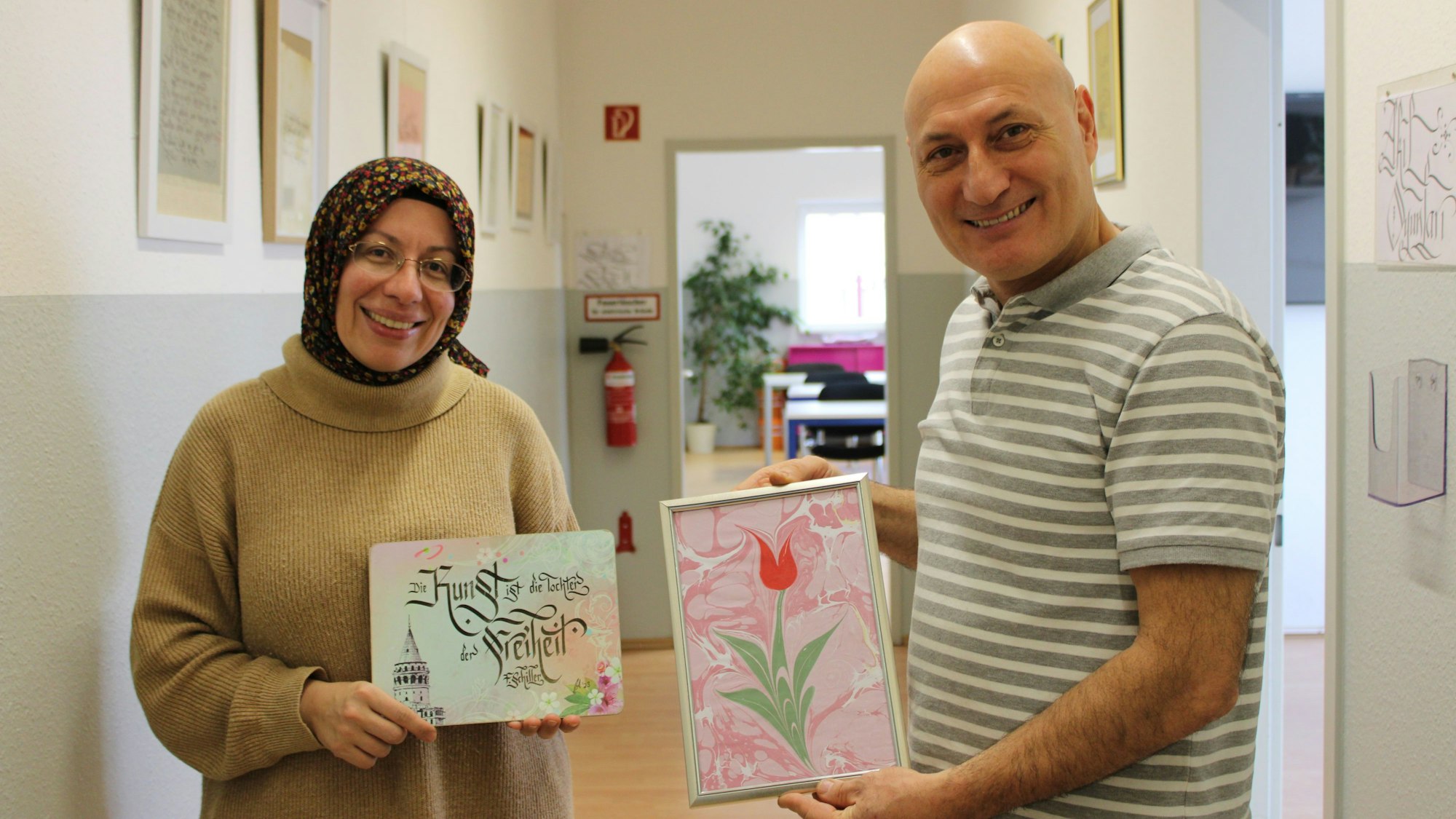 Eine Frau und ein Mann halten zwei Kunstwerke, auf einem ist eine Tulpe zu sehen, auf einem die Aufschrift „Kunst ist die Tochter der Freiheit - F. Schiller“.
