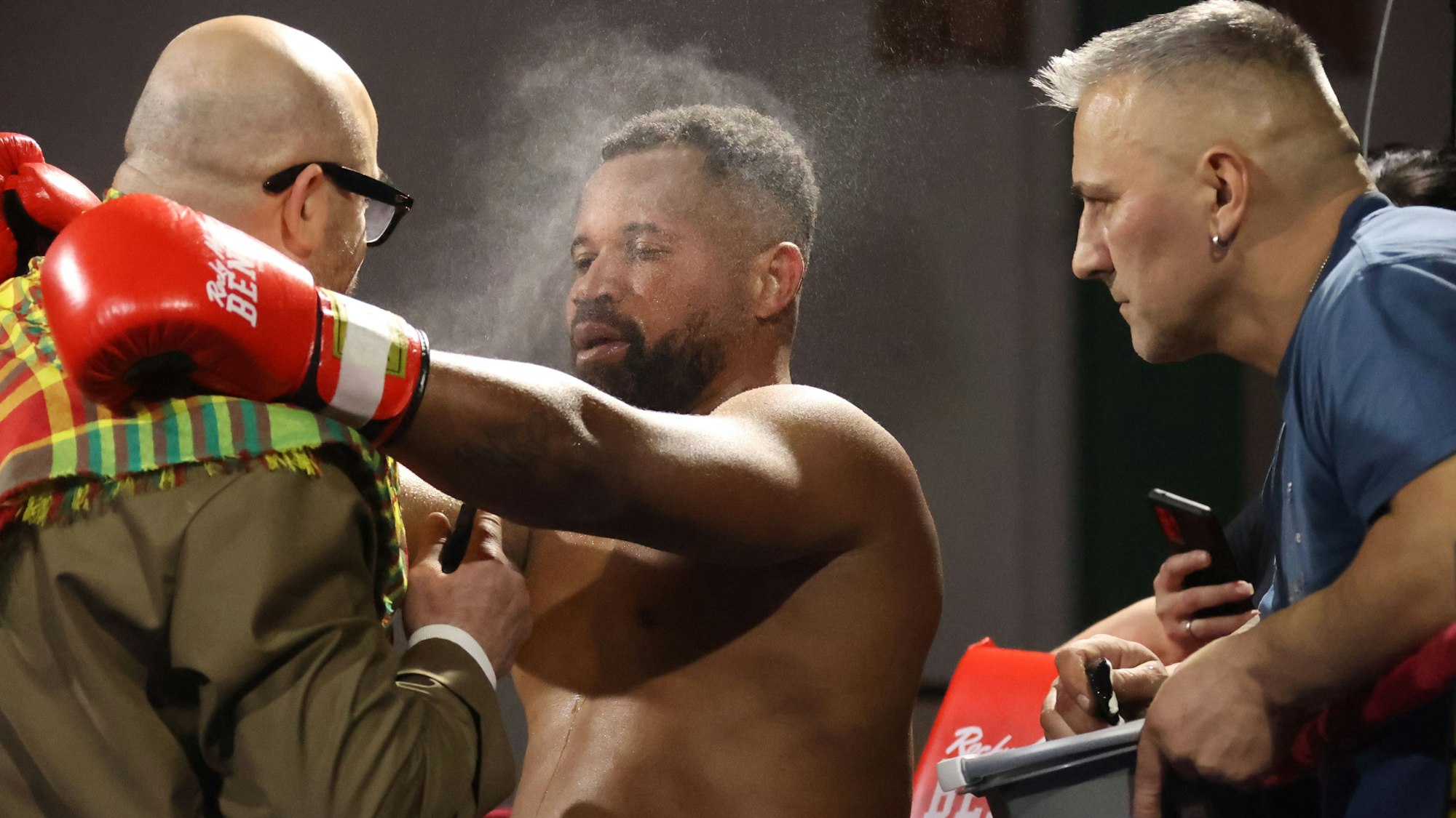 Die beiden Trainer sprechen in der Ringecke mit dem Boxer, der mit Flüssigkeit eingesprüht wird. Der Boxer legt die Arme um die Schulter des Cheftrainers.