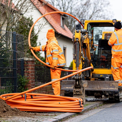 Arbeiter sind mit Bagger und Spaten an einer Straße mit der Erdverlegung von Glasfaserkabeln beschäftigt.