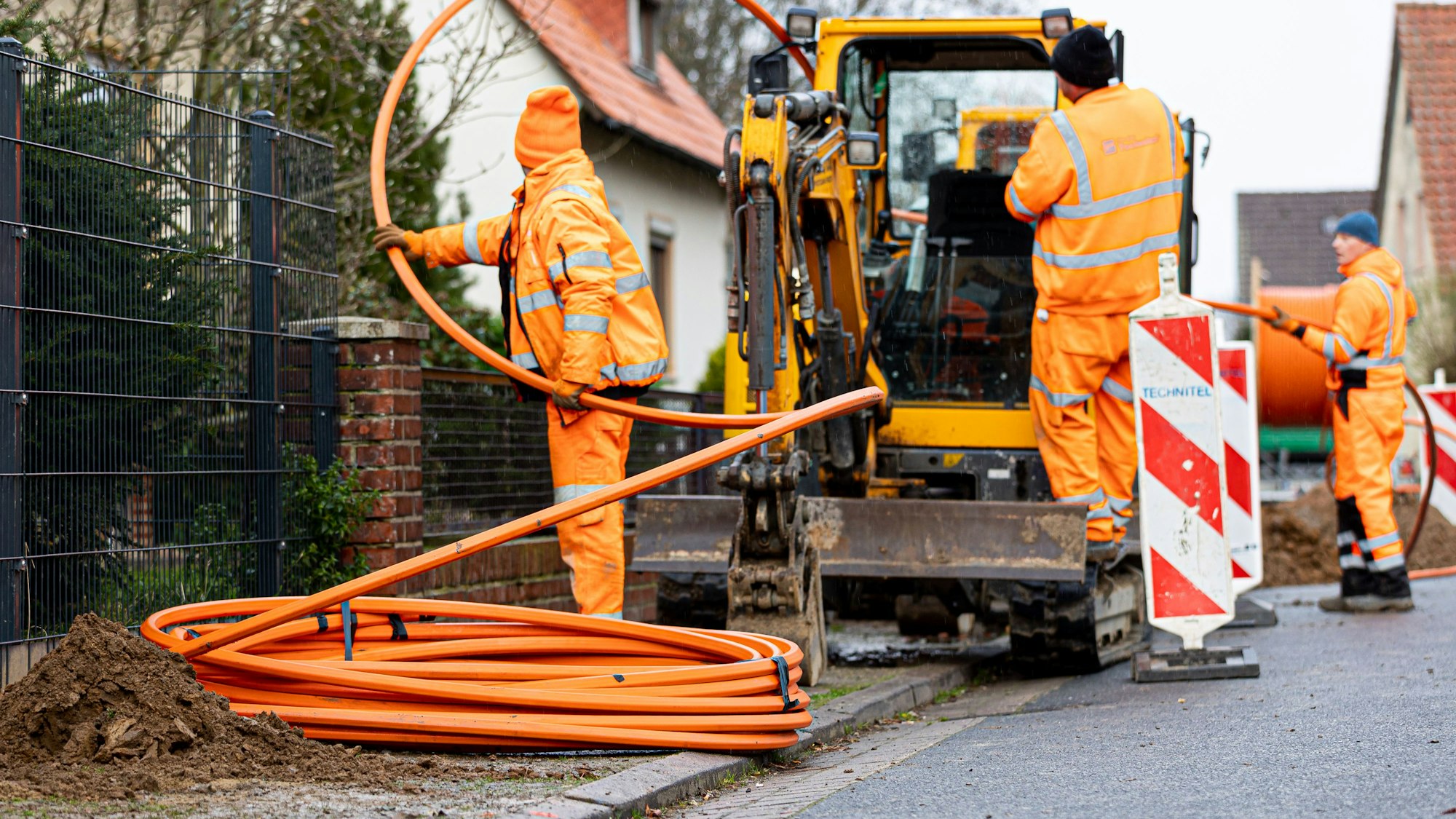 Arbeiter sind mit Bagger und Spaten an einer Straße mit der Erdverlegung von Glasfaserkabeln beschäftigt.