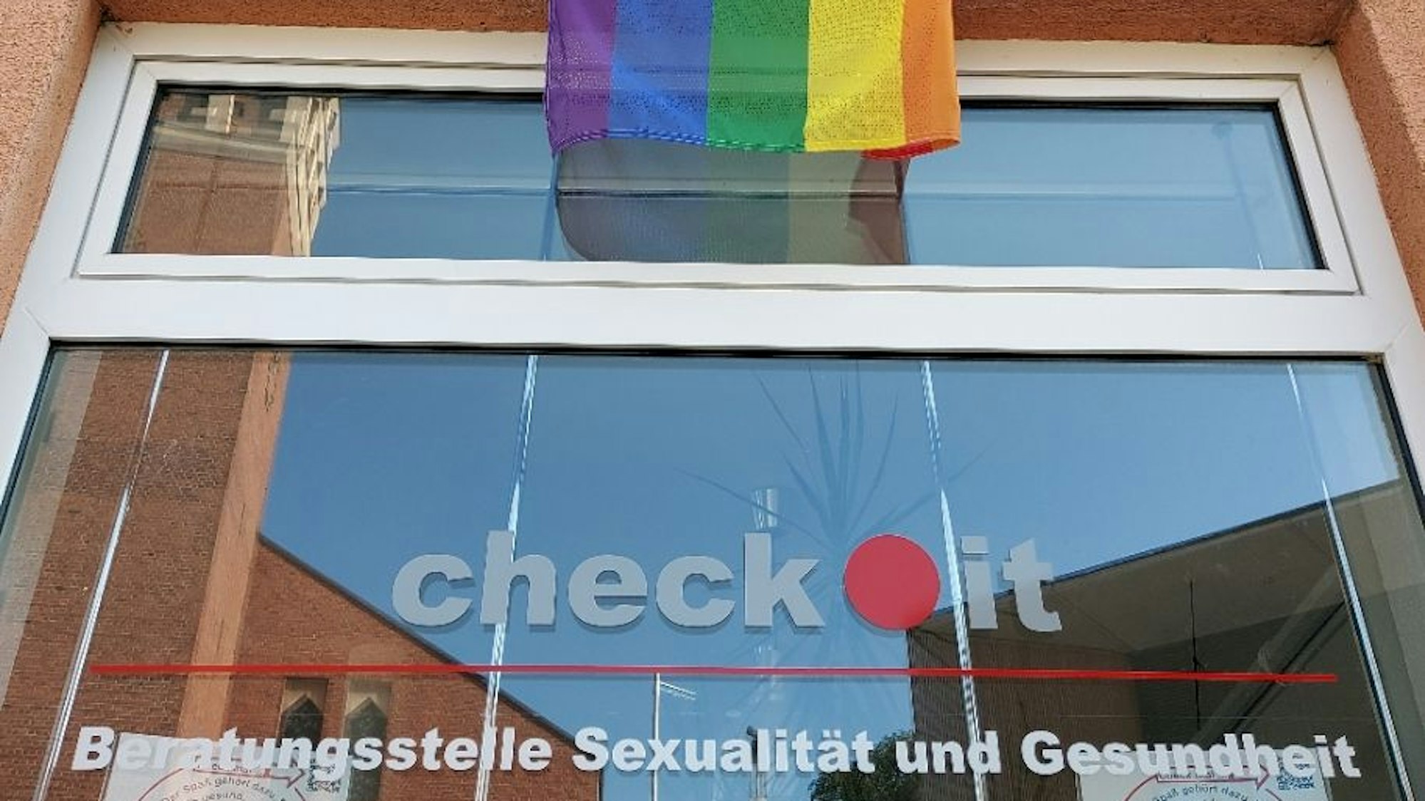 Eine Regenbogenfahne hängt über einem Fenster, darauf ist die Aufschrift "Check.it: Beratungsstelle Sexualität und Gesundheit" zu lesen.