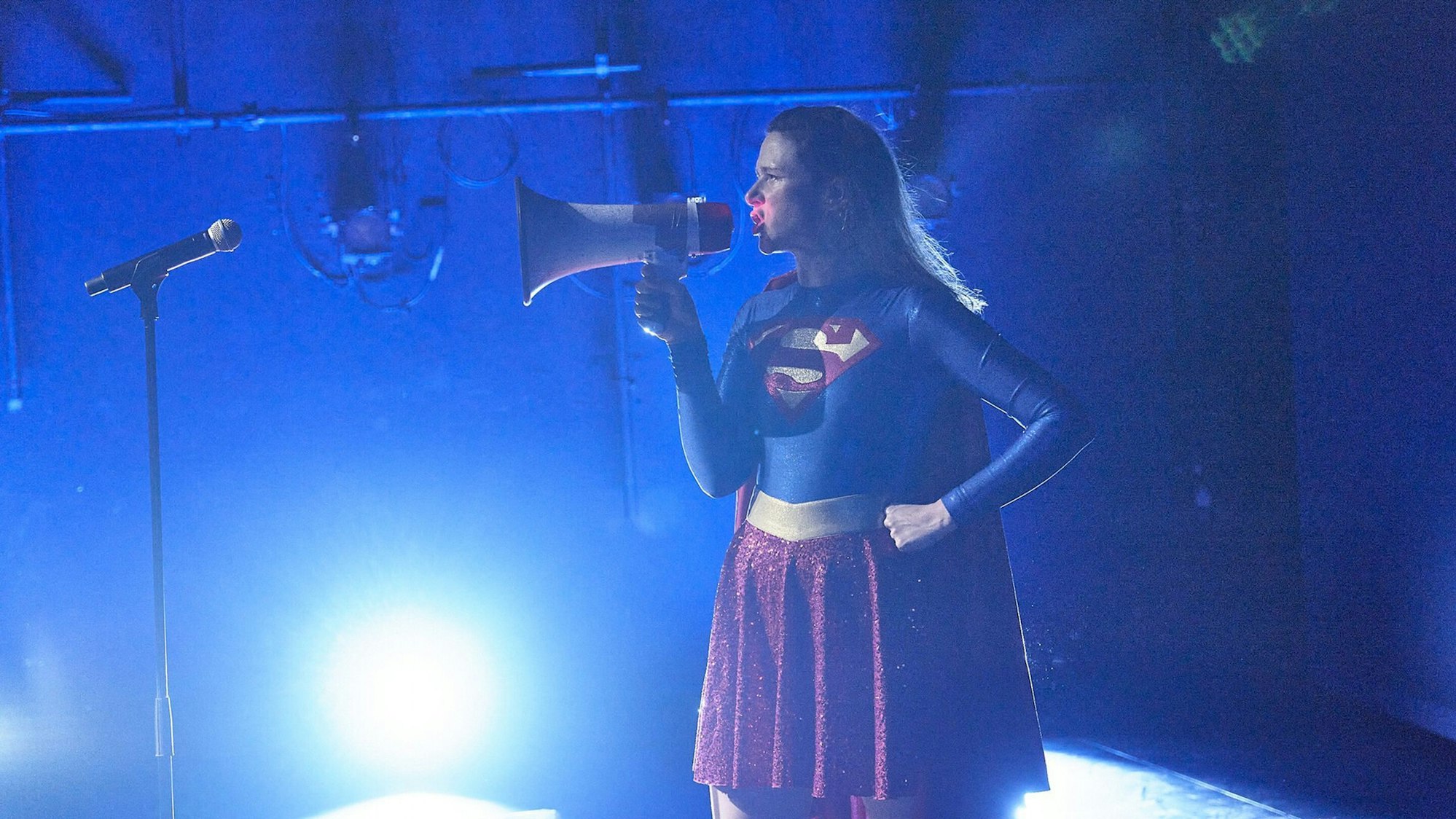 Claude De Demo spricht im Supergirl-Kostüm in ein Megafon.