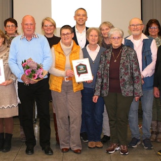 17 Männer und Frauen stehen für ein Gruppenfoto zusammen, in der vorderen Reihe werden Blumensträuße und Urkunden gehalten.