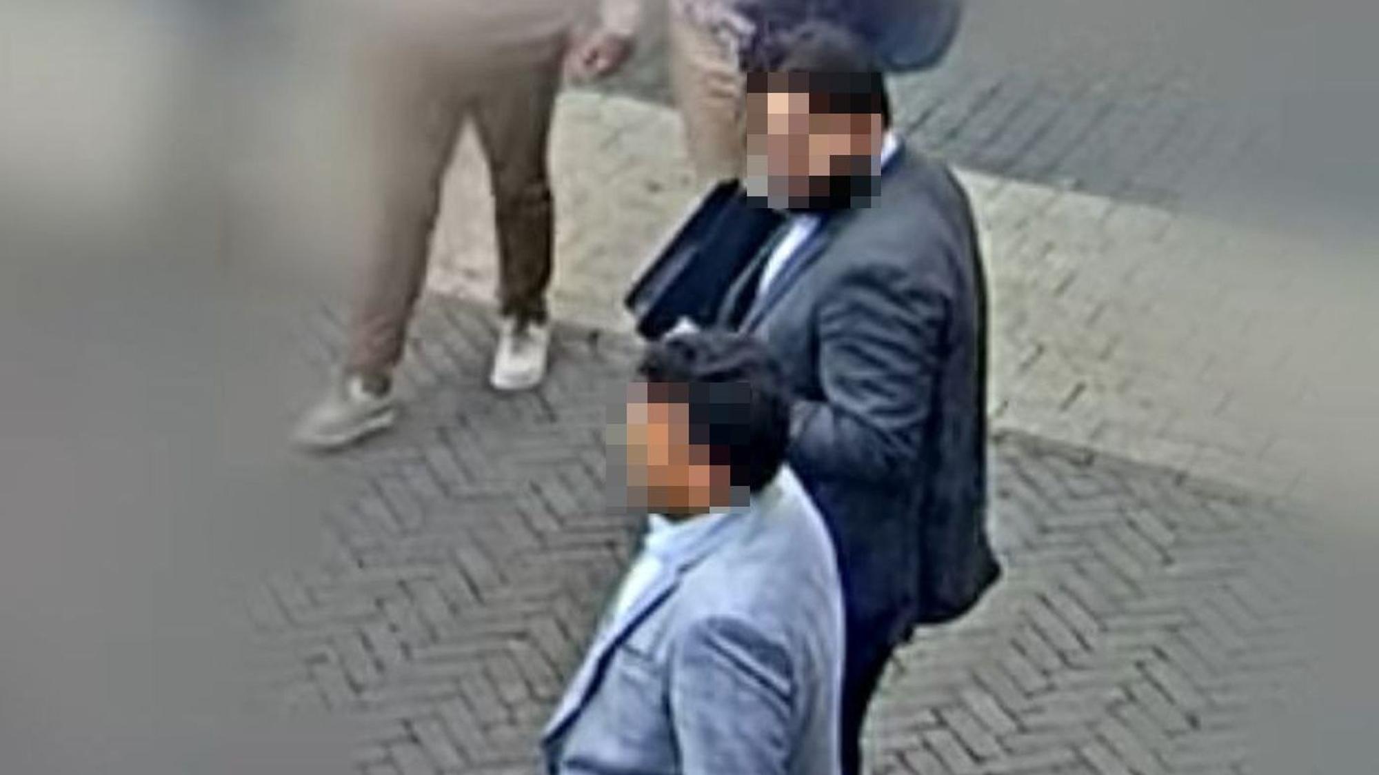 Auf der Aufnahme einer Überwachungskamera sind zwei Männer zu sehen, die Anzüge tragen. Die Gesichter der Männer sind verpixelt.