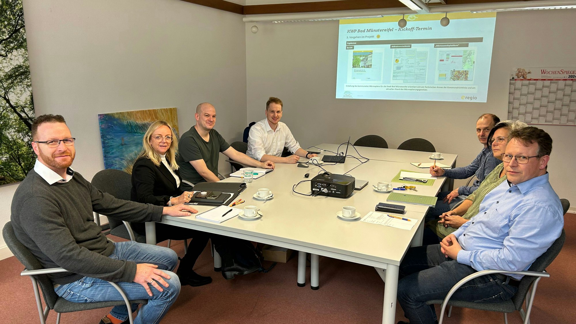 Sieben Menschen sitzen um einen Tisch herum. Per Beamer wird eine Grafik an die Wand geworfen.