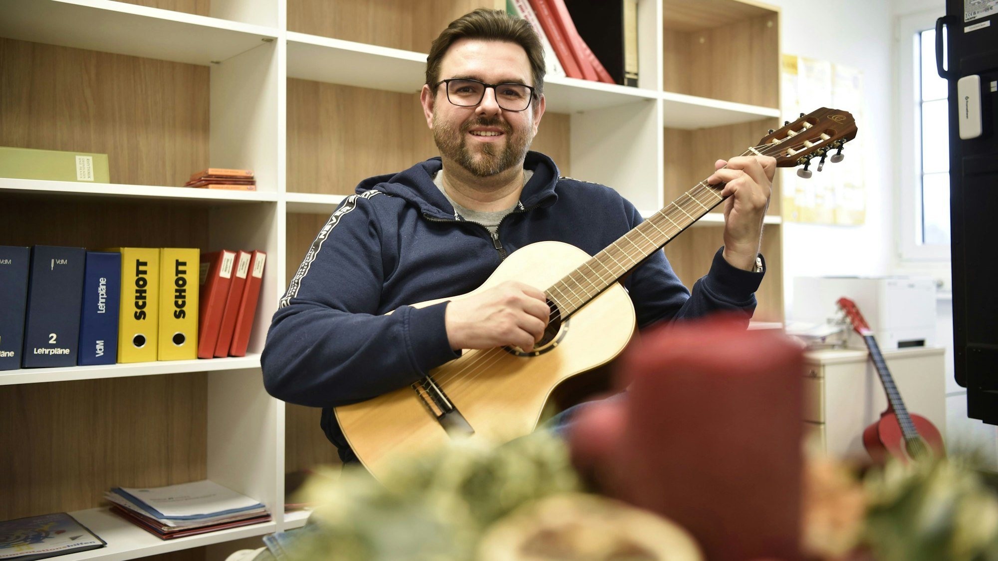 Peter Rack sitzt in seinem Büro und hält eine Gitarre in der Hand. Hinter ihm steht ein Regal, vor ihm eine rote Kerze.