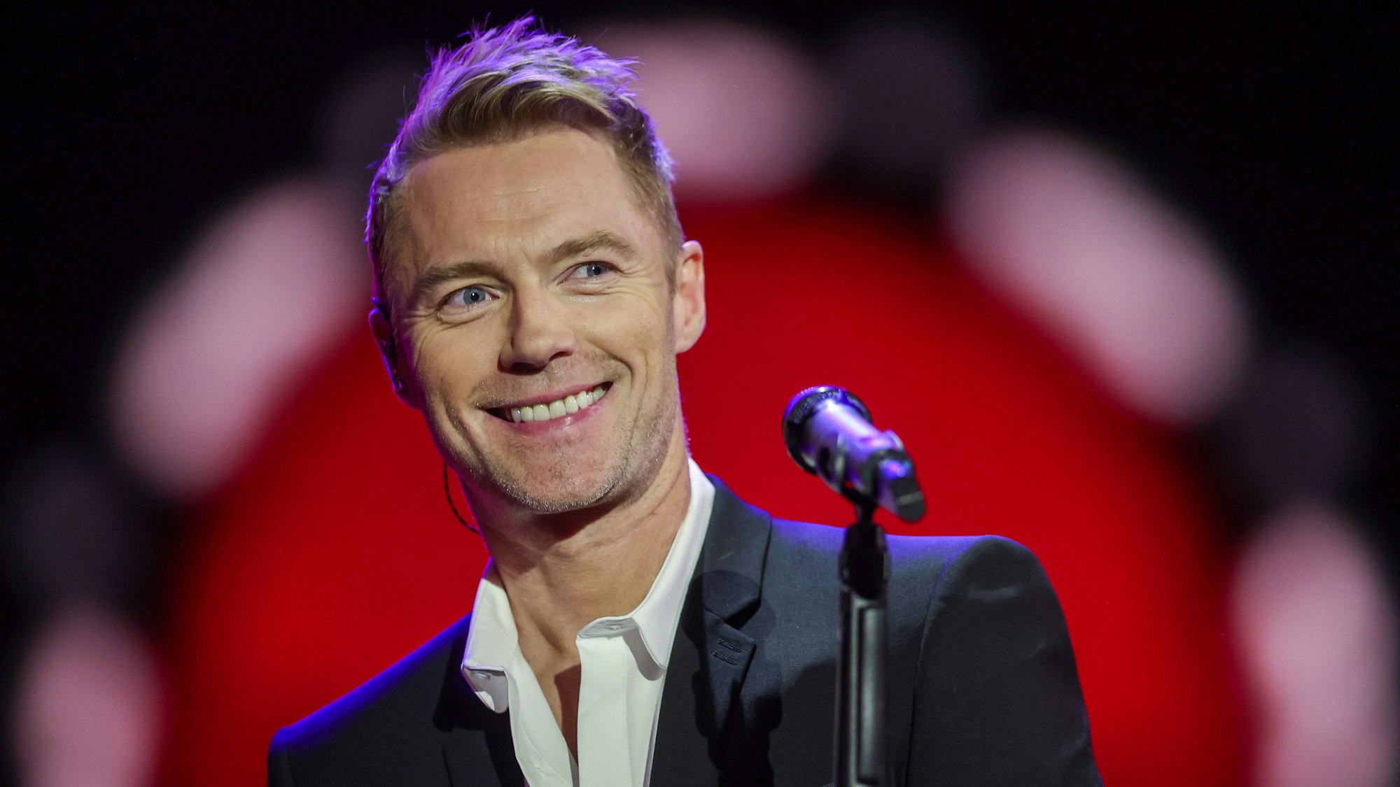 Ronan Keating steht vor einem Mikrofon und lächelt.