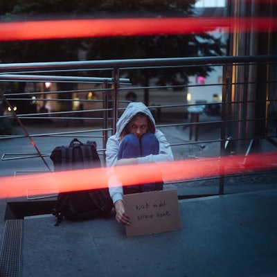 Ein Mädchen mit grauem Kapuzenpullover lehnt in der Nacht an der Gatter eines U-Bahn-Abgangs und hält ein Schild mit der Aufschrift „Weiß nicht wohin heute“ in der Hand.