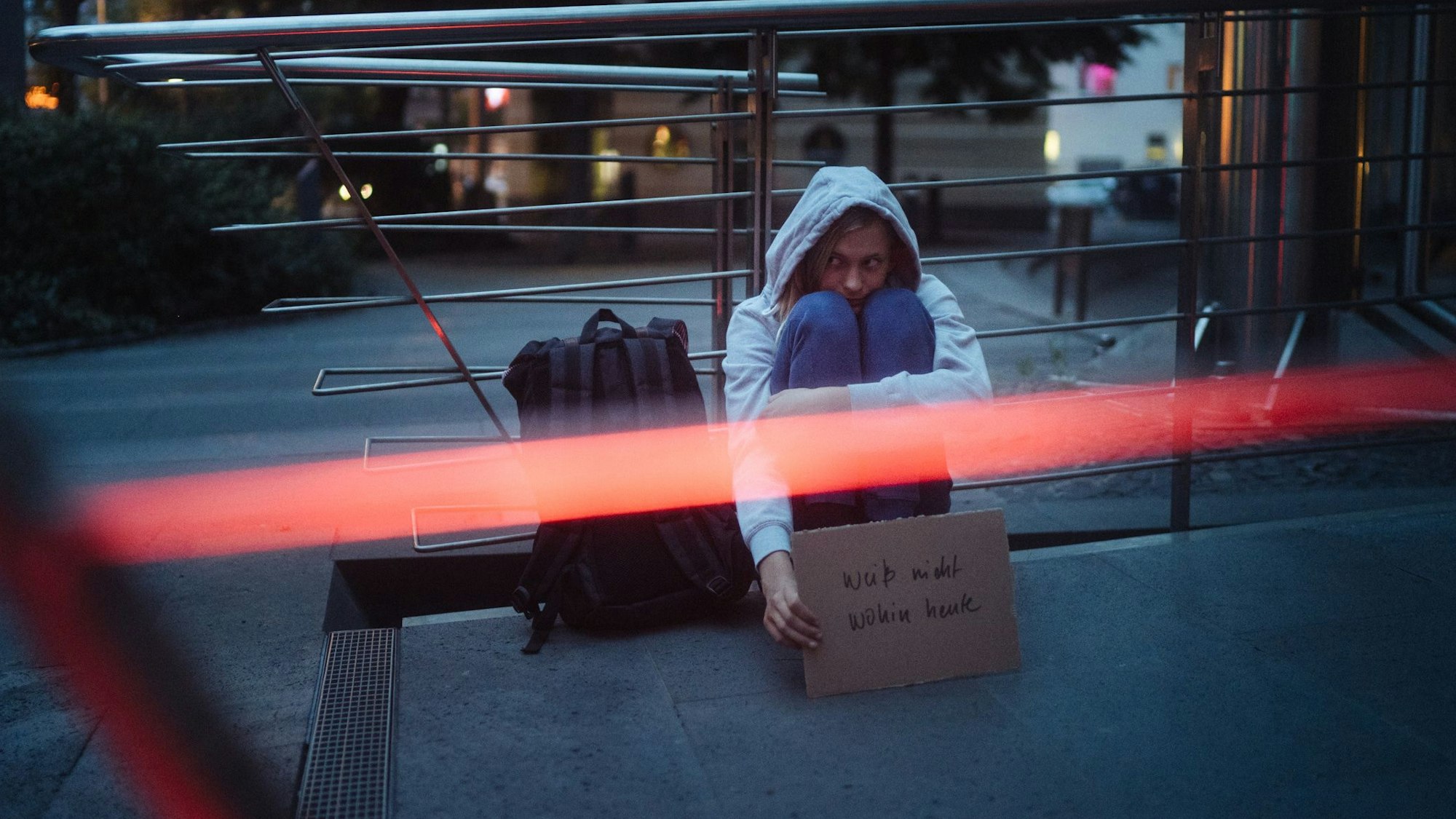 Ein Mädchen mit grauem Kapuzenpullover lehnt in der Nacht an der Gatter eines U-Bahn-Abgangs und hält ein Schild mit der Aufschrift „Weiß nicht wohin heute“ in der Hand.
