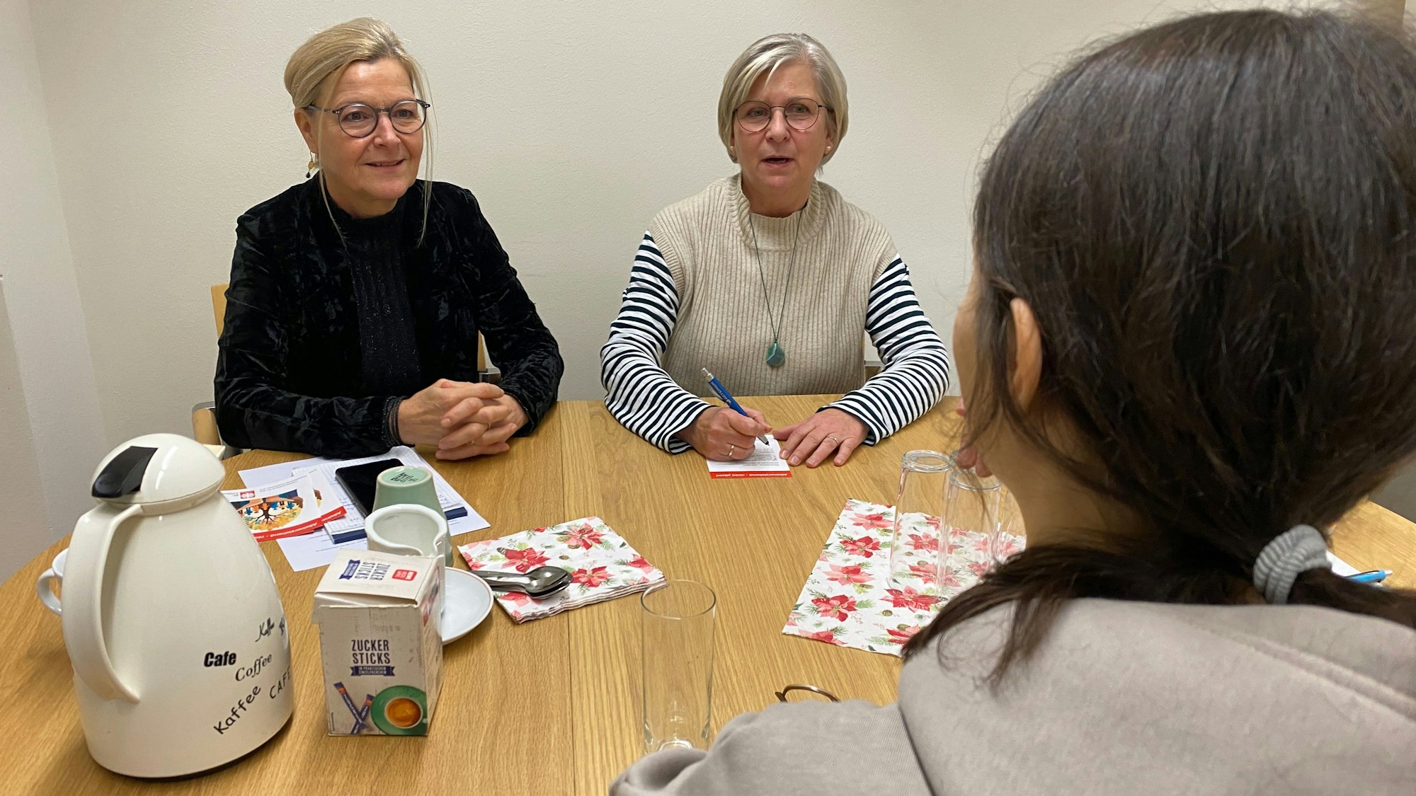 Drei Frauen sitzen an einem Tisch, eine mit dem Rücken zum Betrachter. Auf dem Tisch steht eine Thermoskanne.