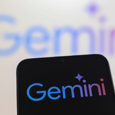 Ein Smartphone mit dem Logo von Gemini vor einer Wand, auf der ebenfalls Gemini steht.
