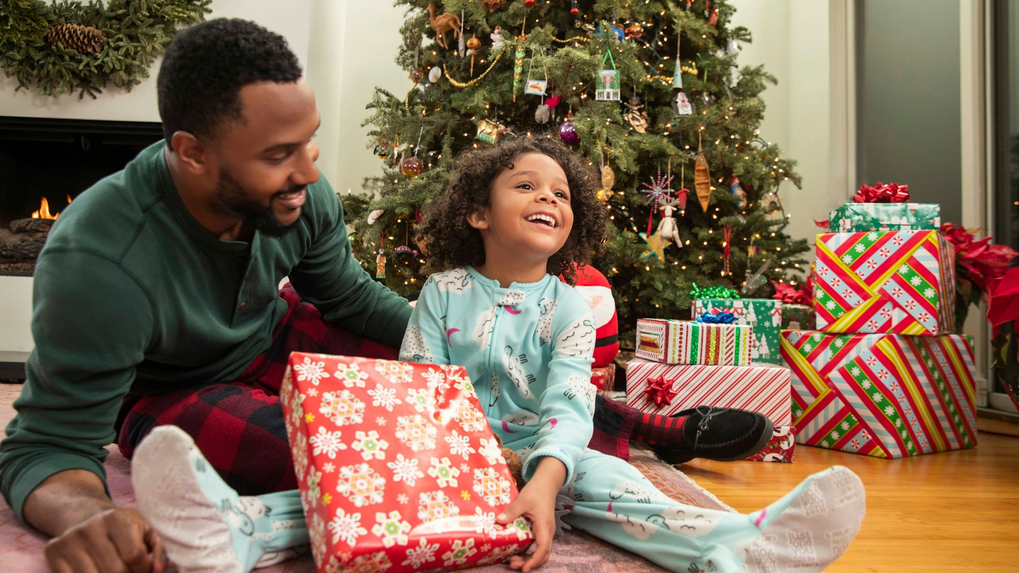 Ein Vater und seine junge Tochter sitzen vor einem Weihnachtsaum, beide strahlen, auf dem Bild sind viele Geschenke zu sehen.