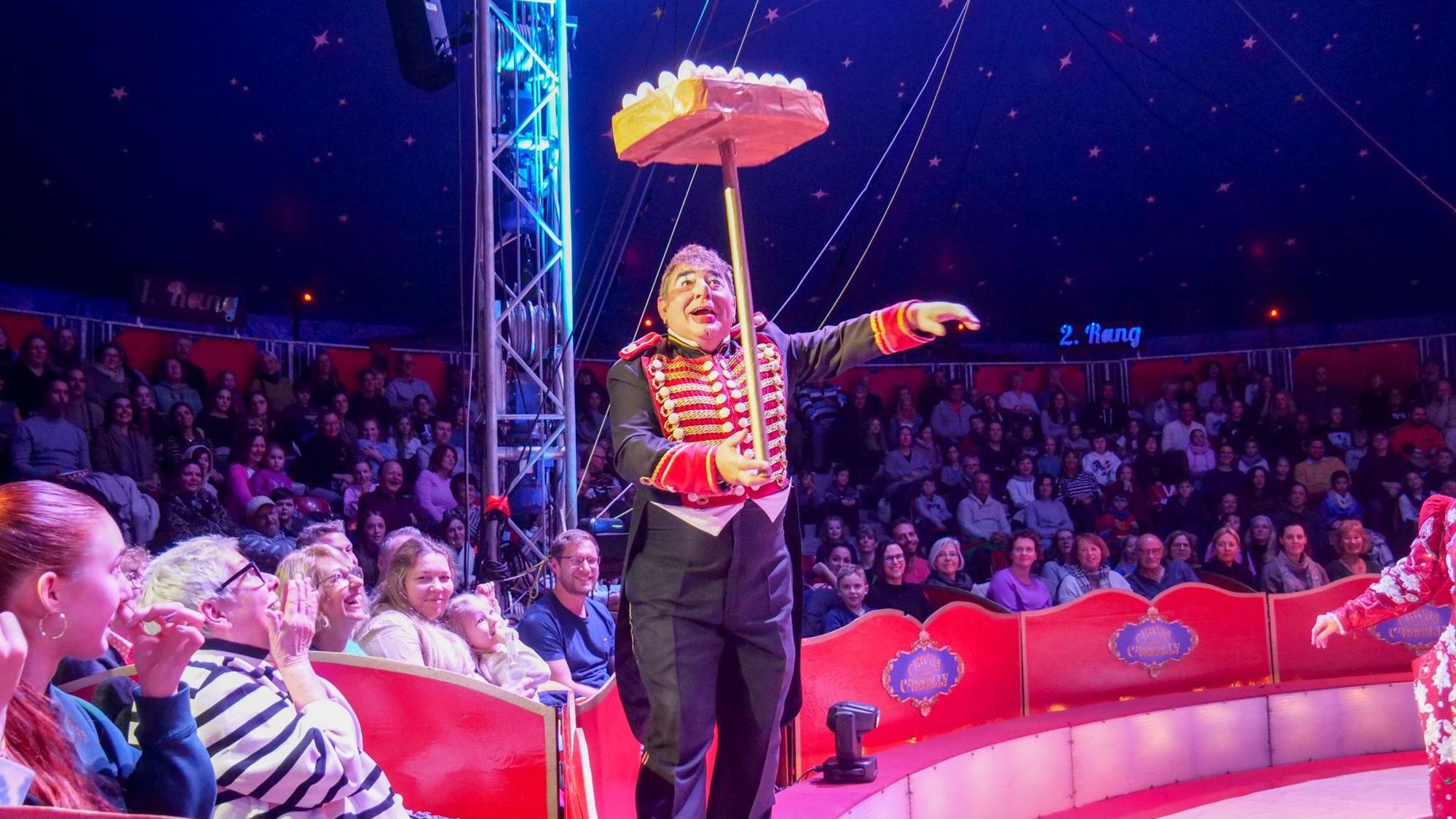 Das Foto zeigt den Clown mit einer Jonglage-Nummer vor den Rängen rund um die Manege.