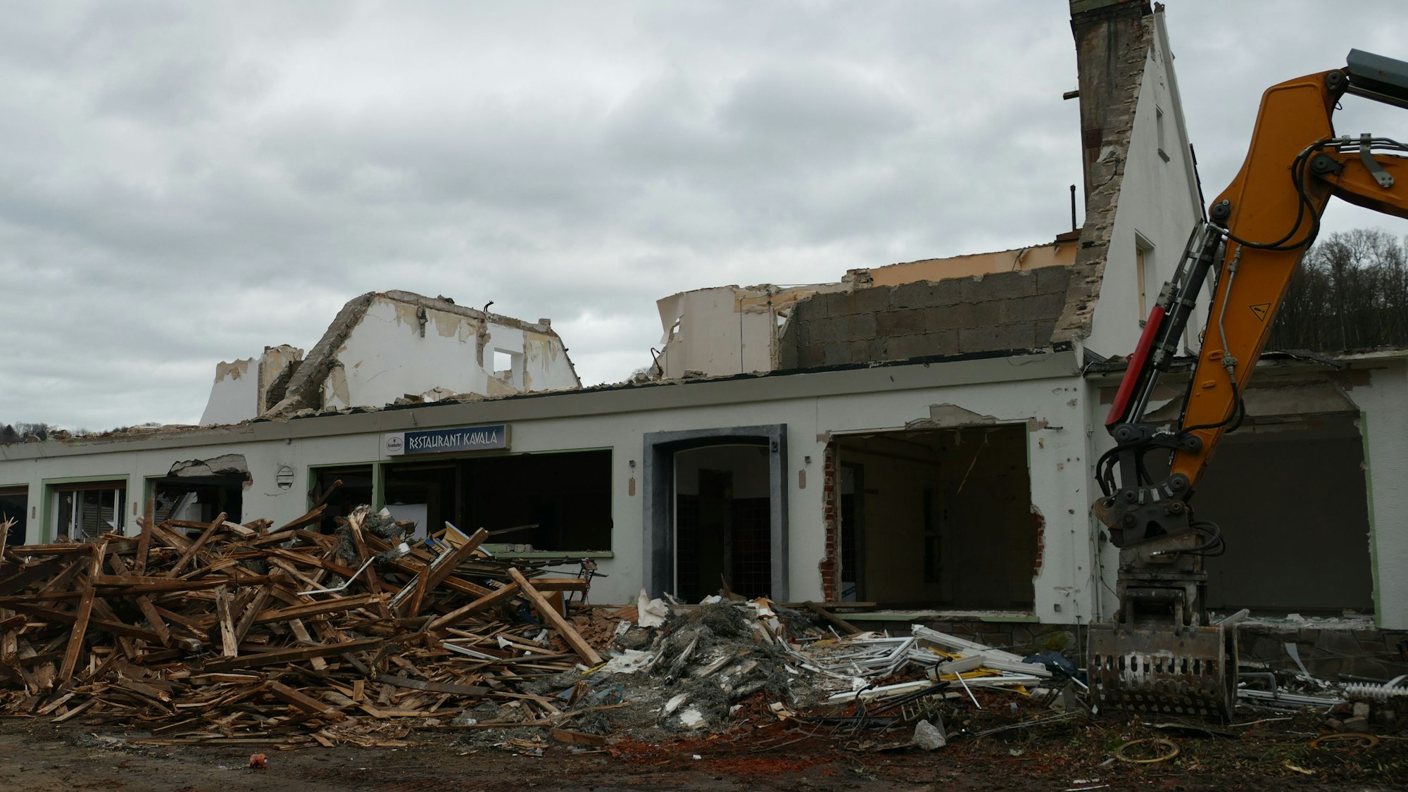 Ein Bagger an einem halb abgerissenen Haus, Balken und Schutt liegen davor.