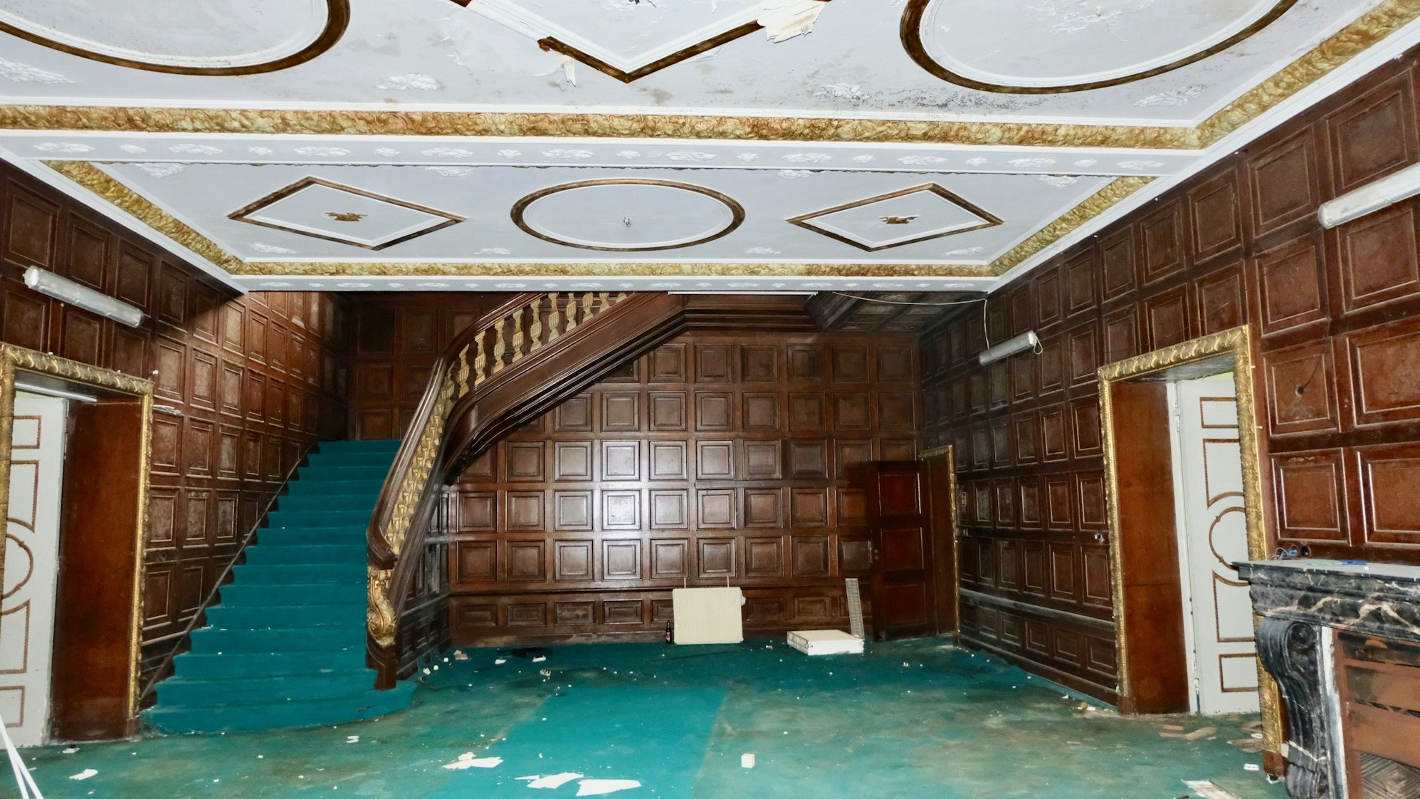 Zu sehen ist eine holzvertäfelte Eingangshalle, von der links und rechts zwei Türen in weitere Zimmer abgehen. Links führt eine Treppe ins Obergeschoss.