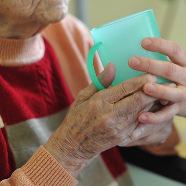 Die Hände einer alten Frau umschließen eine grüne Plastiktasse, sie werden von der Hand einer jungen Frau gestützt.