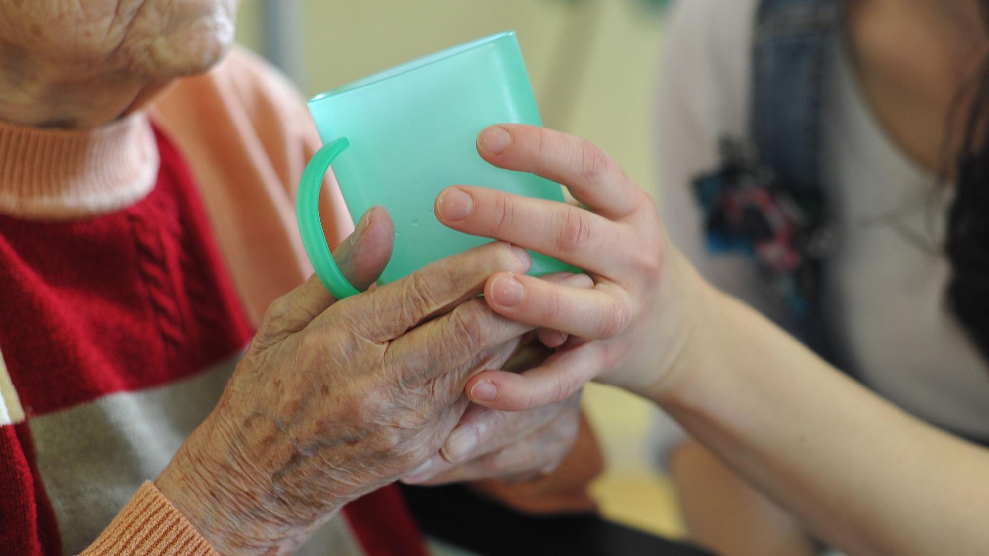 Die Hände einer alten Frau umschließen eine grüne Plastiktasse, sie werden von der Hand einer jungen Frau gestützt.