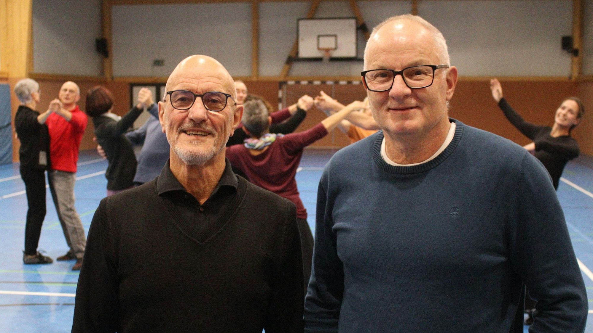 Zwei Männer stehen nebeneinander und blicken in die Kamera. Im Hintergrund tanzen Paare in einer Sporthalle.