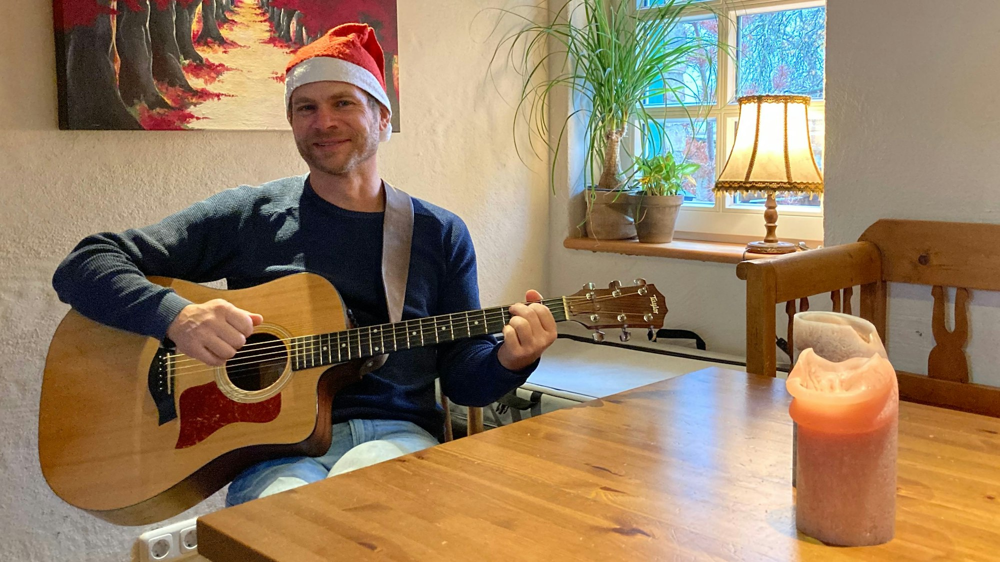 Der Kindermusiker Uwe Reetz spielt sitzend am Tisch Gitarre, während er eine Weihnachtsmütze trägt.