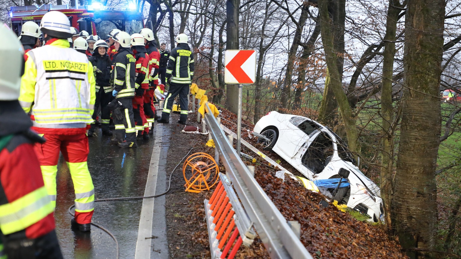 Ein schwer beschädigtes Auto steht vor einem Baum in einer Böschung, auf der Straße oberhalb stehen Feuerwehrleute.