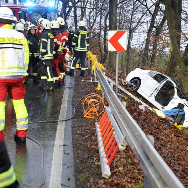 Ein schwer beschädigtes Auto steht vor einem Baum in einer Böschung, auf der Straße oberhalb stehen Feuerwehrleute.