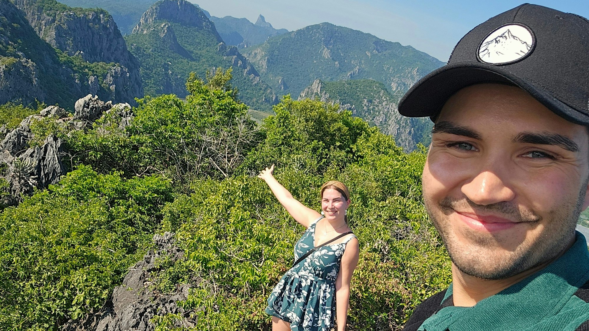 Ein junger Mann schießt ein Selfie, im Hintergrund weist eine junge Frau in eine tropische Gebirgslandschaft.