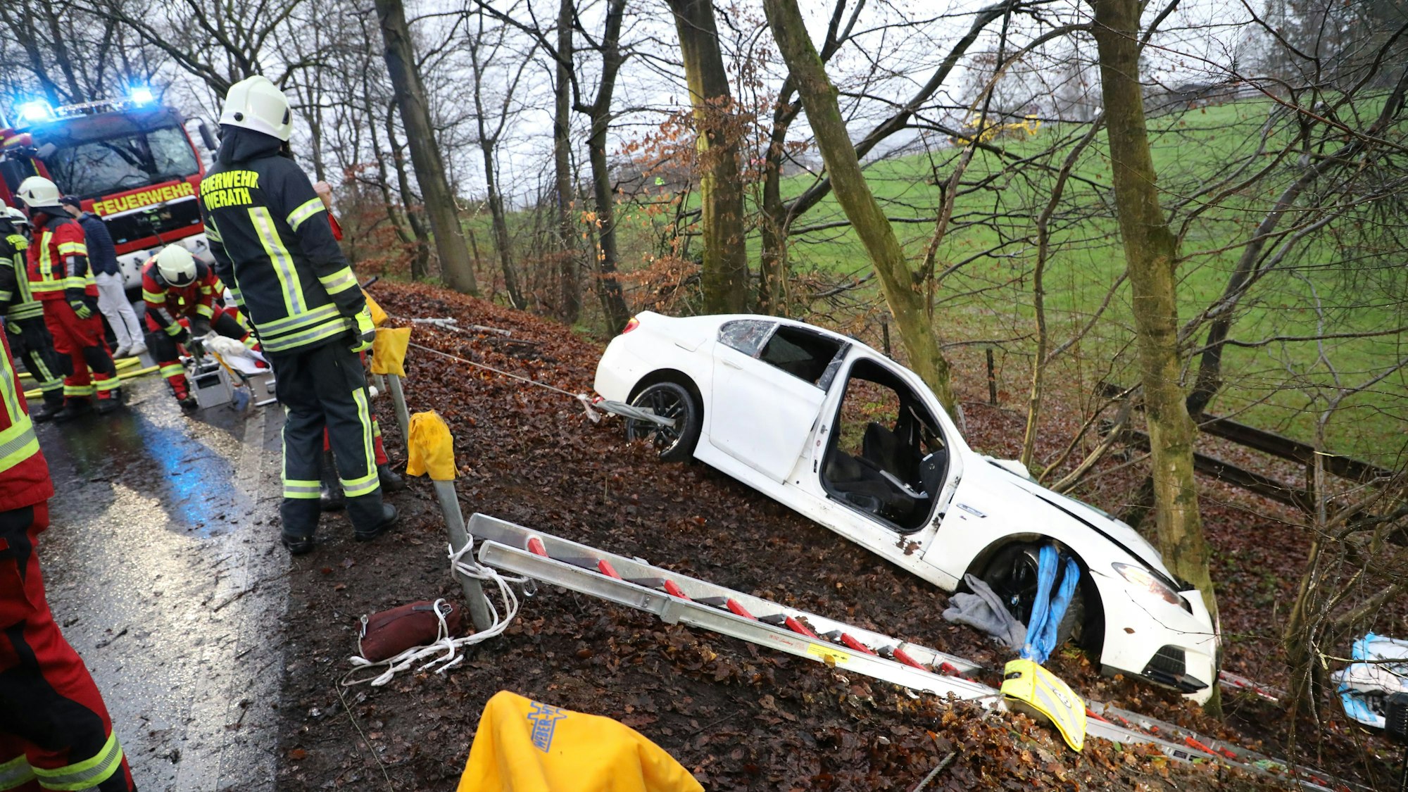 Ein schwer beschädigtes Auto steht vor einem Baum in einer Böschung, auf der Straße oberhalb stehen Feuerwehrleute.