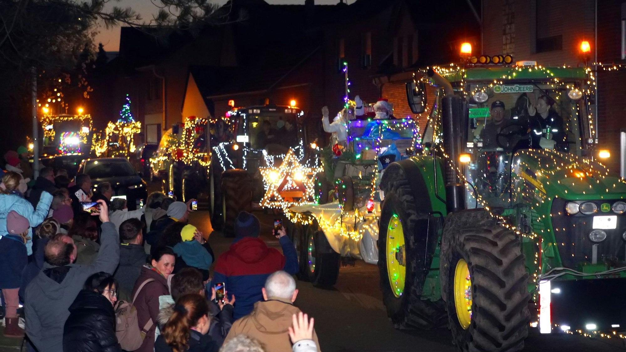 Menschen stehen an Straßenrand und bestaunen mit Lichtern geschmückte Traktoren, die vorbei fahren.