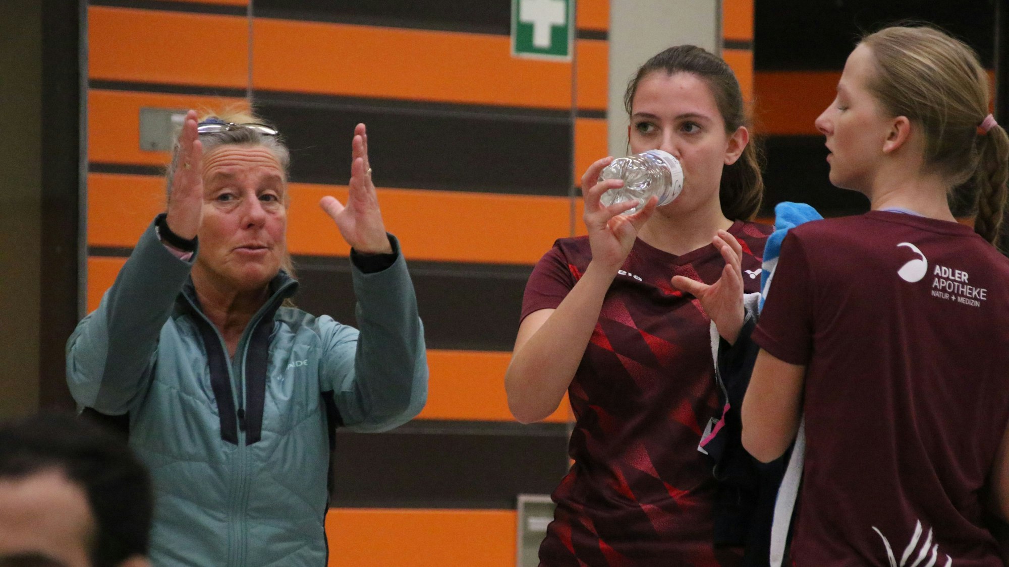 Kalls Badminton-Trainerin Iris Schröder redet gestenreich mit zwei Spielerinnen, von denen eine aus einer Wasserflasche trinkt.