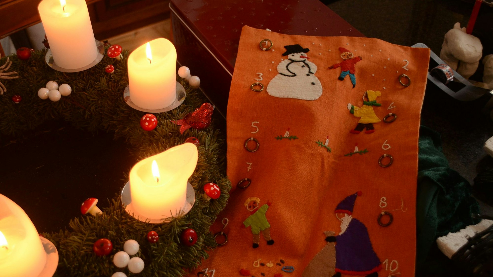 Ein Adventskalender liegt auf dem Tisch neben einem Adventskranz, an dem die Kerzen brennen.