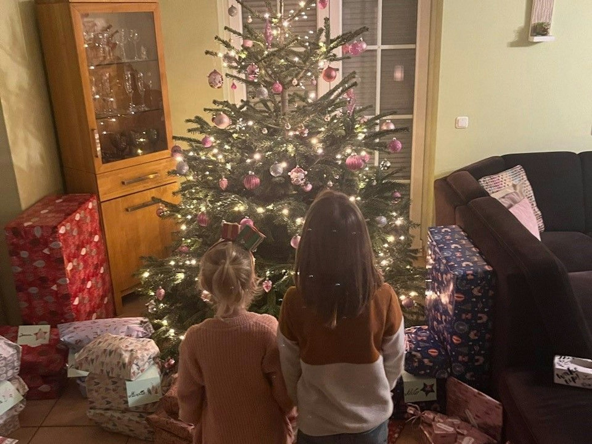Zwei Kinder stehen vor einem geschmückten Weihnachtsbaum. Drumherum sind zahlreiche verpackte Geschenke.