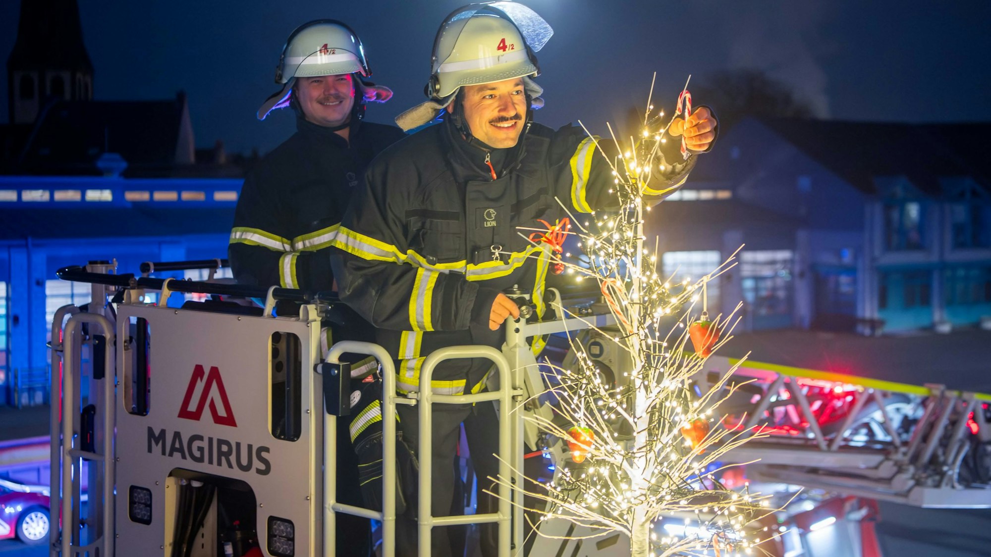 Das Bild zeigt zwei Feuerwehrmänner im Korb der Drehleiter. Sie schmücken einen kleinen, beleuchteten Weihnachtsbaum.