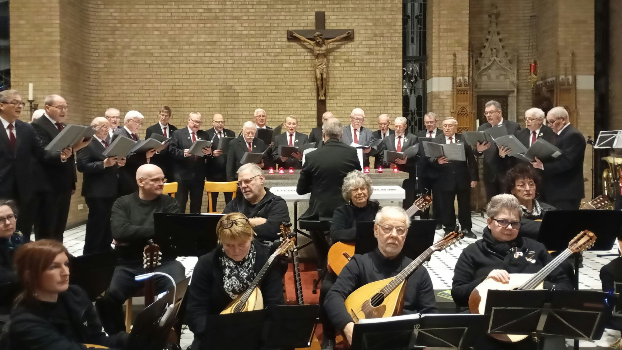 Auf dem Bild ist das Mandolinenorchester zu sehen, die Musiker sitzen und tragen schwarze Kleidung. Hinter ihnen steht der Männerchor in einem Halbkreis.
