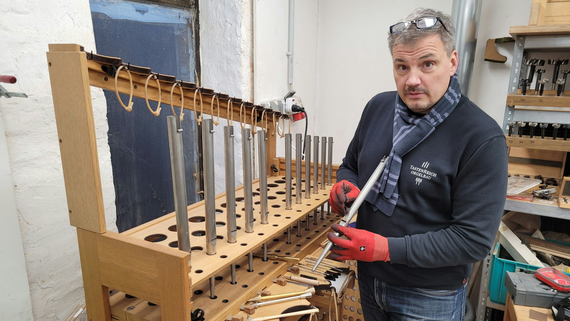Der Orgelbauer in seiner Werkstatt, er steht vor einem Gestell mit mehreren Pfeifen.