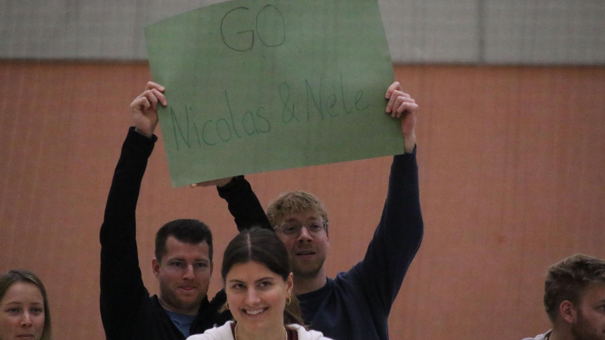 Mit einem grünen Pappschild feuern zwei Männer einen Spieler und eine Spielerin an.