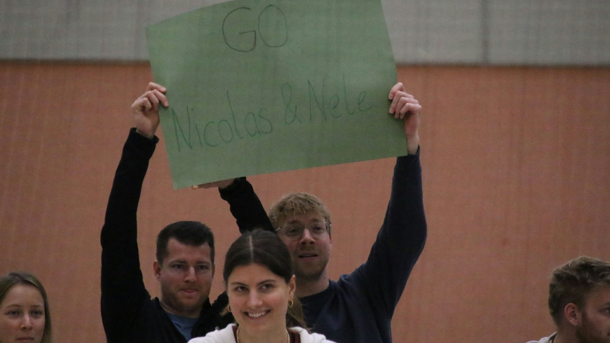 Mit einem grünen Pappschild feuern zwei Männer einen Spieler und eine Spielerin an.