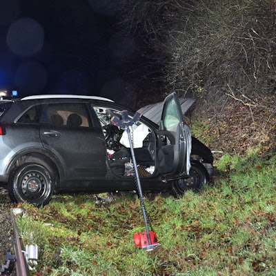 Ein verbeultes Auto steht neben einer regennassen Straße, im Hintergrund Einsatzfahrzeuge von Polizei und Feuerwehr.