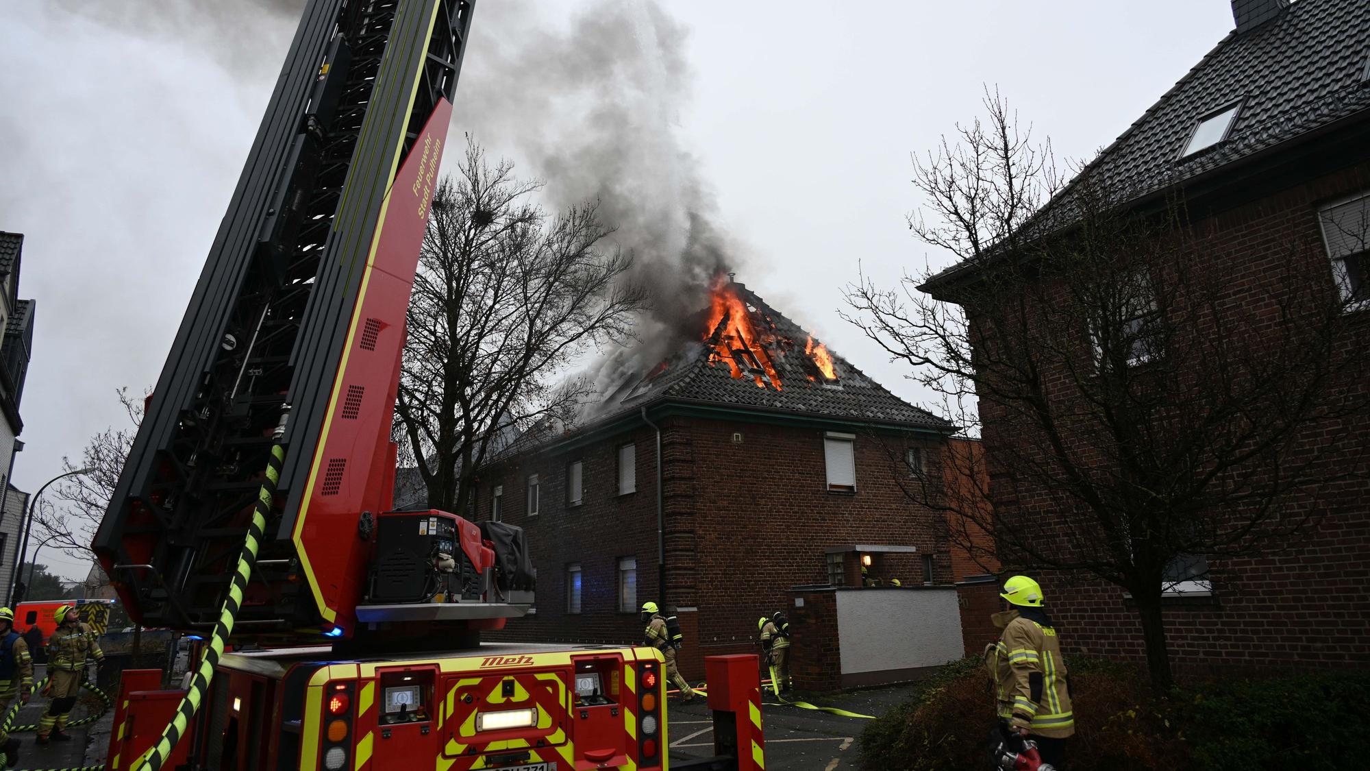 Ein Dachstuhl brennt im Hintergrund, ein Fahrzeug der Feuerwehr und Feuerwehrleute stehen im Vordergrund.