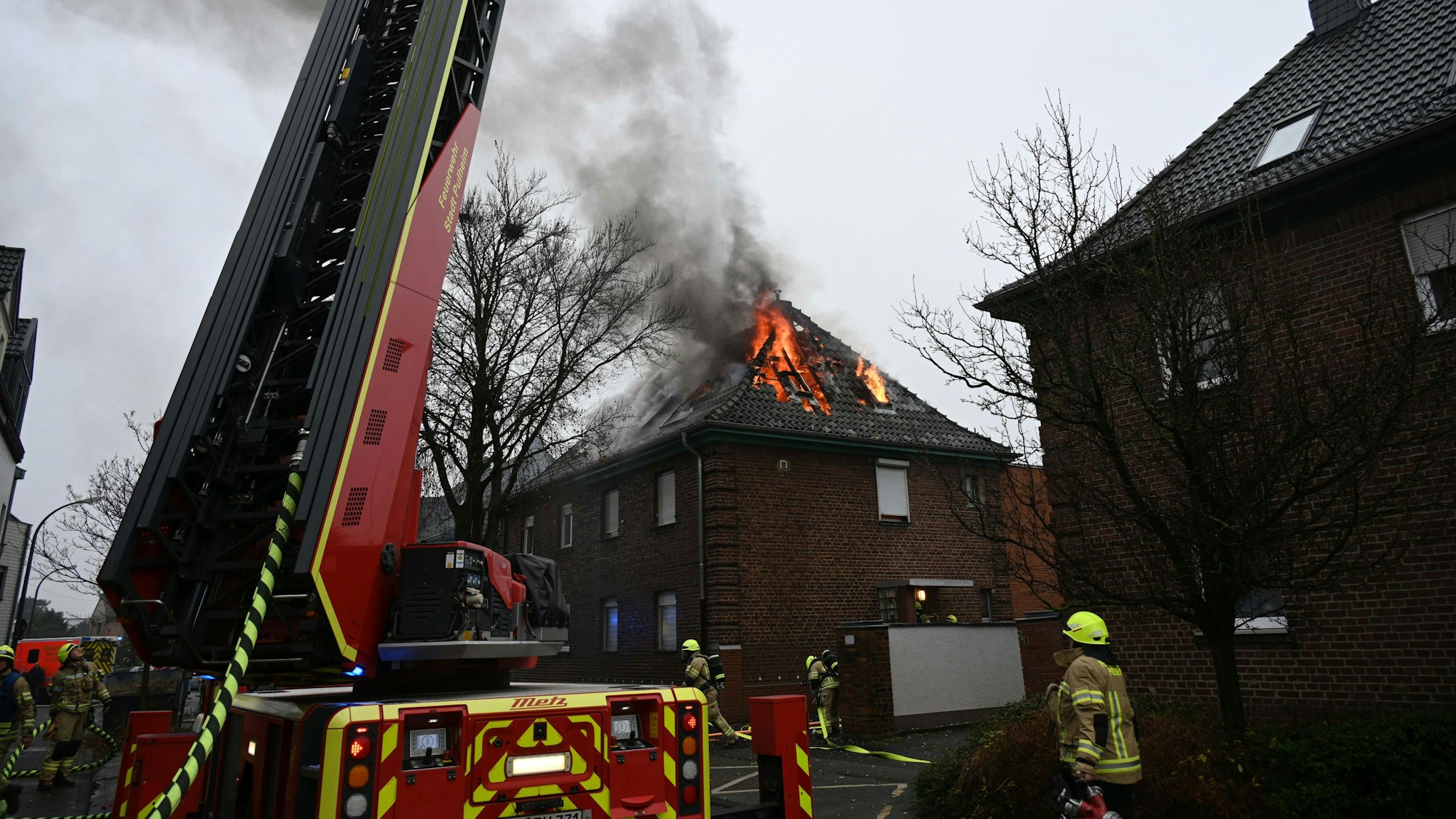 Ein Dachstuhl brennt im Hintergrund, ein Fahrzeug der Feuerwehr und Feuerwehrleute stehen im Vordergrund.