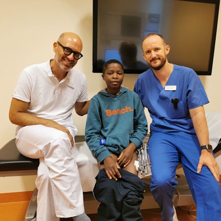 Zwei Ärzte sitzen mit einem dunkelhäutigen Jungen auf einer Behandlungsliege in einem Krankenhaus