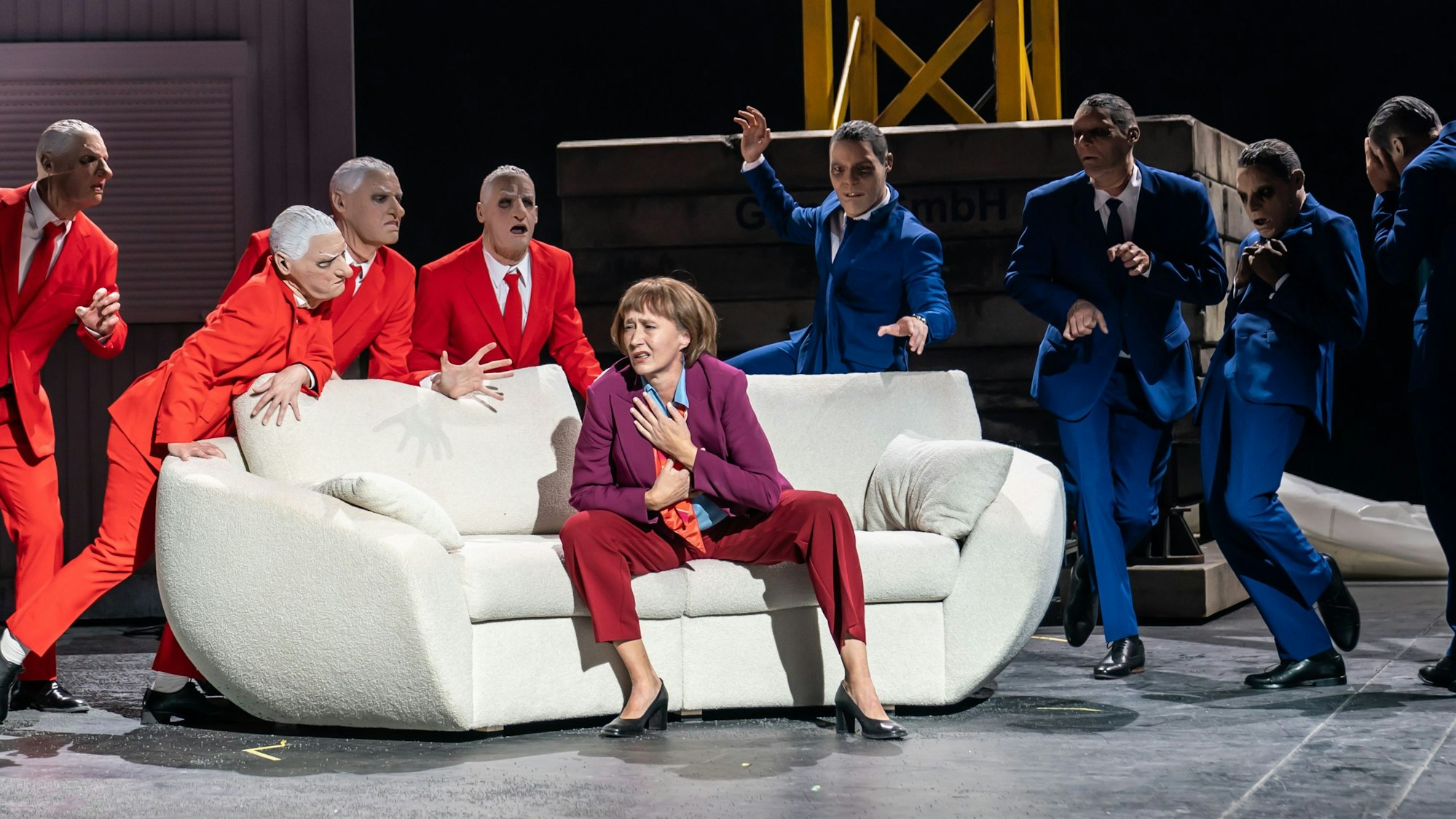 Anja Laïs sitzt in ihrer Rolle als Kölner Oberbürgermeisterin auf einem weißen Sofa im Depot 1 des Kölner Schauspiels, umringt von gestikulierenden Herren in orangen und blauen Anzügen.