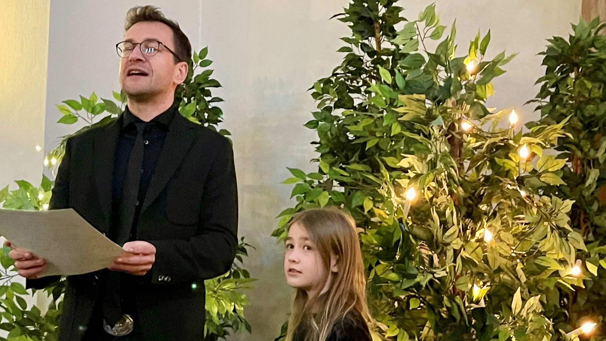 Ein Mann mit Brille zitiert einen Text, er hält ein Papier in den Händen, neben ihm steht ein langhaariges Mädchen.