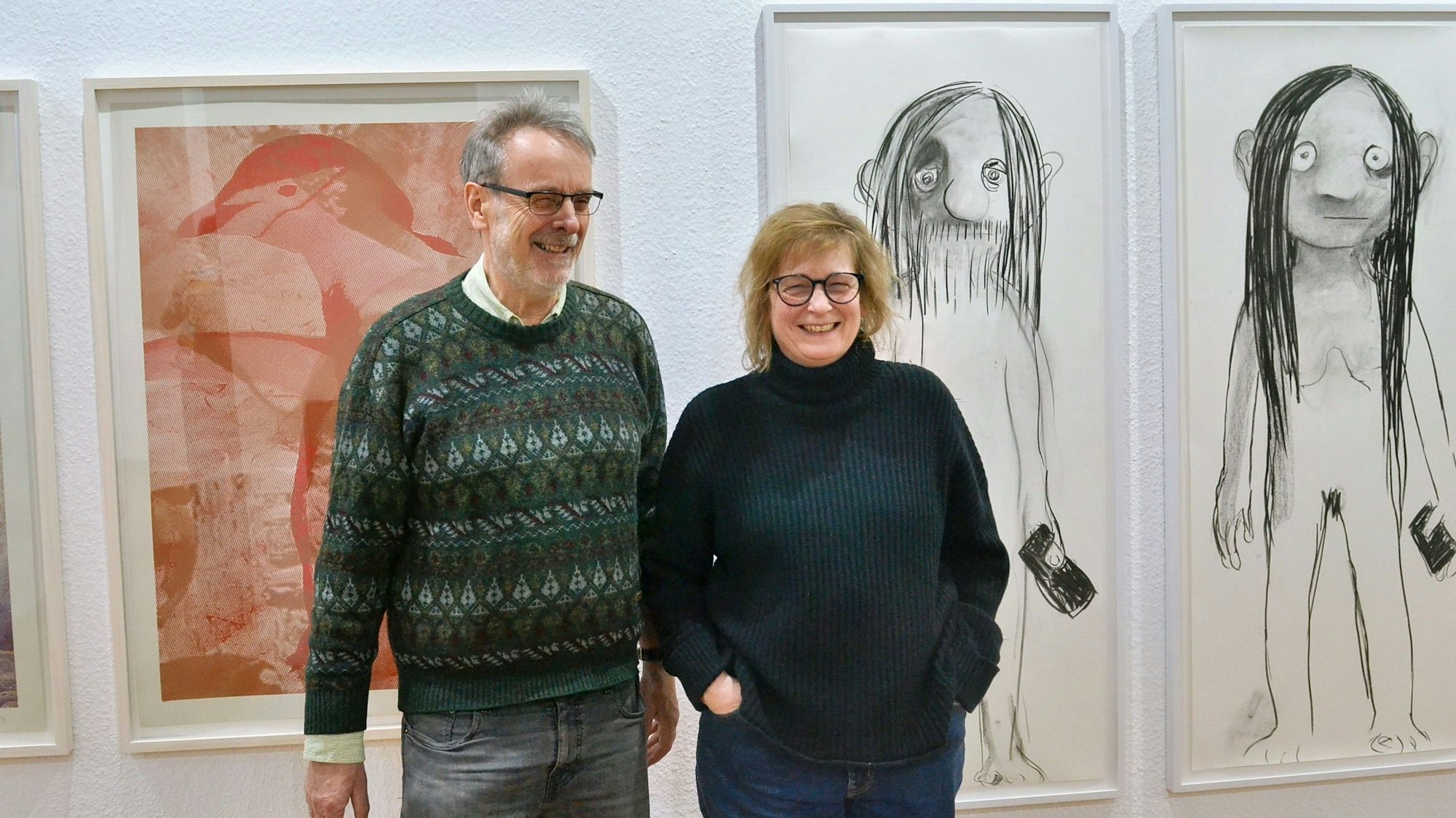 Heike Kati Barath und Paul Schwer stehen in der Galerie Roy in Zülpich vor drei gerahmten Kunstwerken, die an einer weißen Wand hängen.