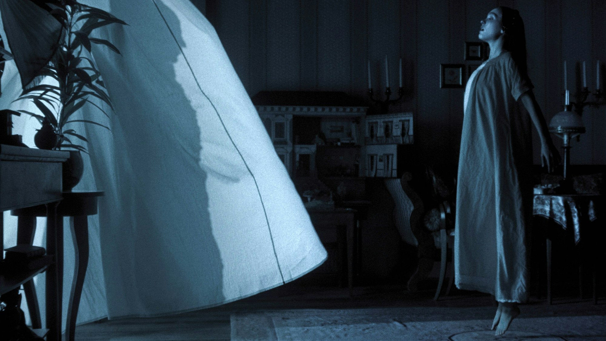 Lily-Rose Depp scheint in einer blau getünchten Szene über dem Boden zu schweben. Im wehenden Vorhang des offenen Fensters erkennt man die Silhouette des Vampirs Nosferatu.