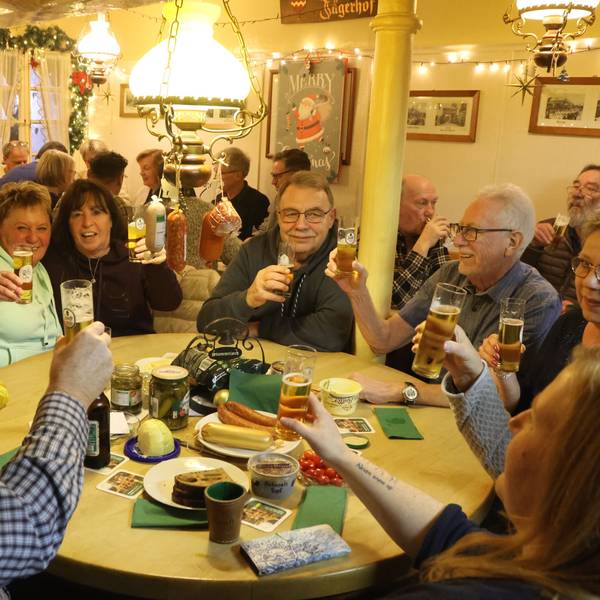 Zu sehen sind Menschen, die in einer Kneipe an einem Tisch ein Würfelspiel spielen. An der Lampe hängen Würste.